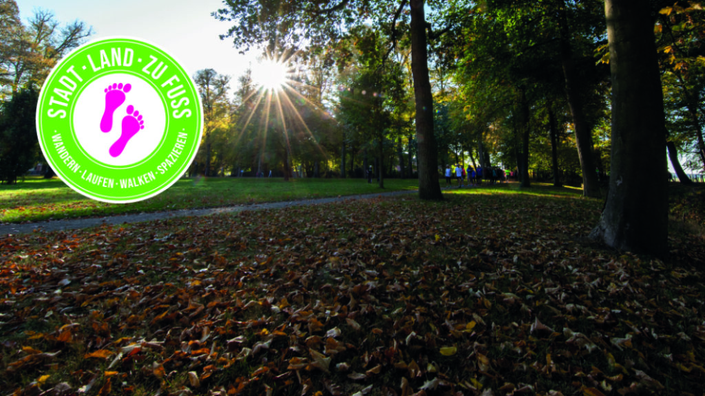 Das Bild zeigt im Hintergrund einen Park im Herbst, in dem Spaziergänger*innen unterwegs sind. Im Vordergrund ist das Logo der Aktion Stadt Land zu Fuß zu sehen.