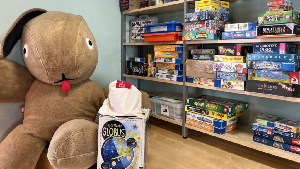 Auf der linken Seite des Bildes sitzt ein großer Stofftierhase, rechts davon sind Regale voll mit Brettspielen