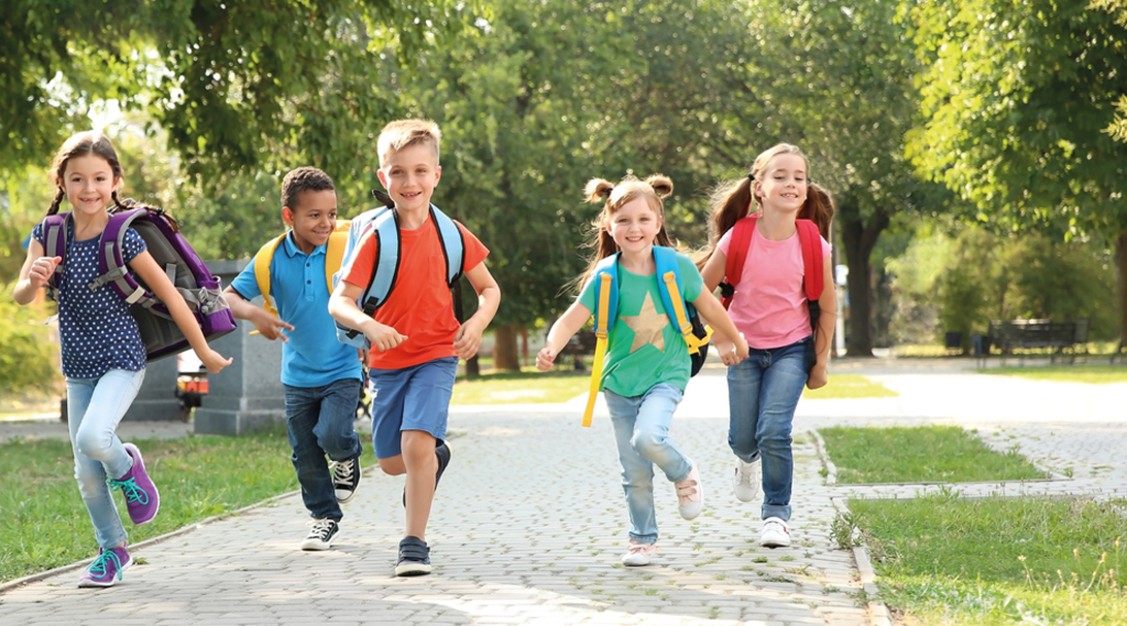 Fünf Kinder mit Schulranzen auf dem Rücken laufen durch einen Park Richtung Schule.