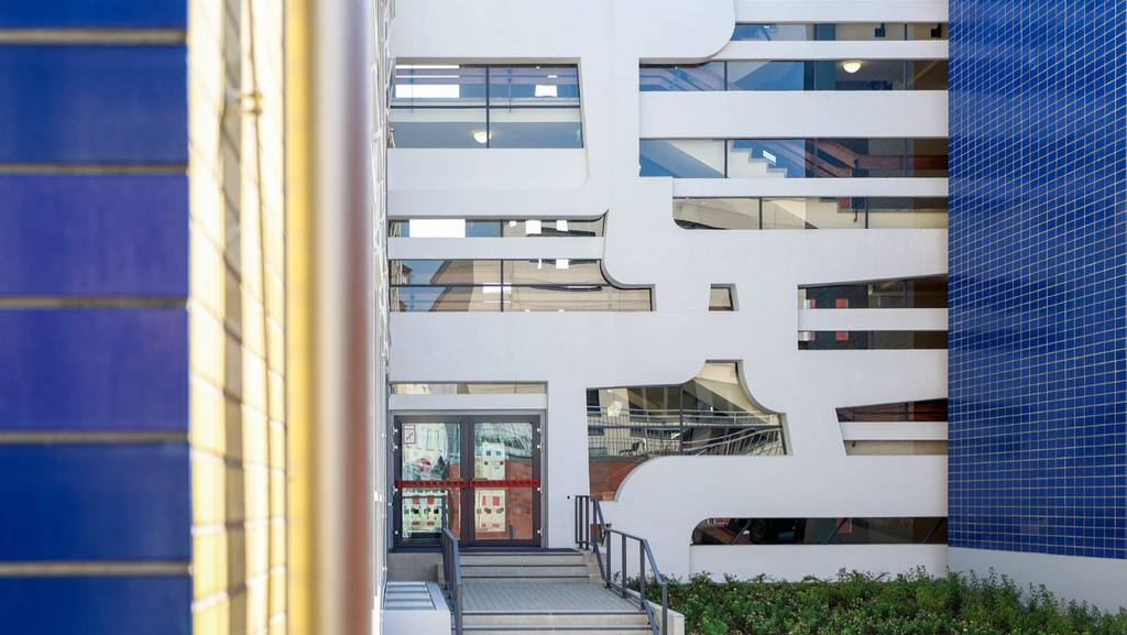 Eine Treppenhaus-Fassade von außen mit weiß verputzten geschwungenen Betonanteilen und vielen, kunstvoll eingesetzten Glasscheiben neben weiteren Fassadenteilen mit blauen Wandfliesen.