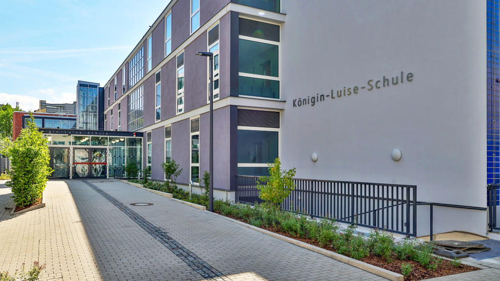 Ein gepflasterter Weg mit begrünten Beeten links und rechts führt zu dem im Schatten gelegenen Haupteingang der Schule. An der Fassade ist der Schriftzug „Königin-Luise-Schule“ zu lesen. 