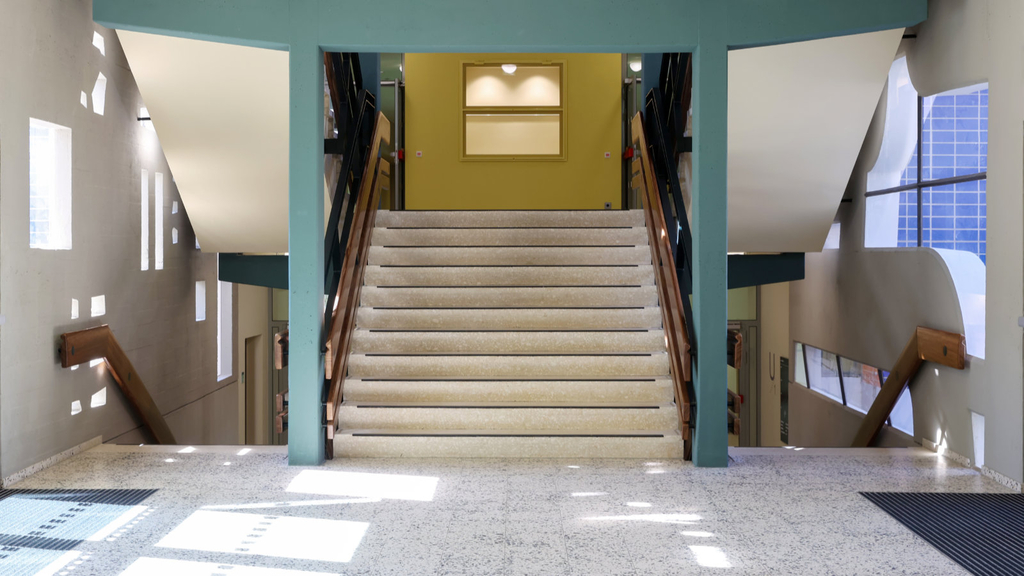 Durch Öffnungen in einer Glasbetonfassade fällt Sonnenlicht auf die Zwischenebene eines Treppenhauses mit Steinböden, Holzgeländern und farblich abgesetzten Pfeilern, Stützen und Wandbereichen. 