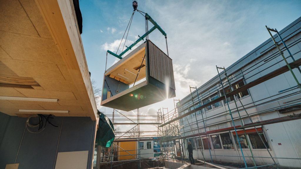 Ein Holzmodul am Kranausleger in der Luft hängend wird in Richtung seines künftigen Platzes auf ein Fundament bewegt.