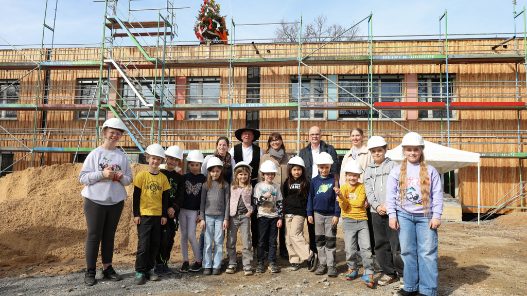 Schüler*innen mit Schulleiterin Jacqueline Fletcher (Grundschule Am Rosenmaar), Bauleiter Thomas Ristau (Blumer Lehmann AG), Petra Rinnenburger (Technische Betriebsleitung der Gebäudewirtschaft), Markus Greitemann (Baudezernent der Stadt Köln) und Gesine Appel (Architektin baut Architektur) vor dem Rohbau aus Holzmodulen mit Richtbaum