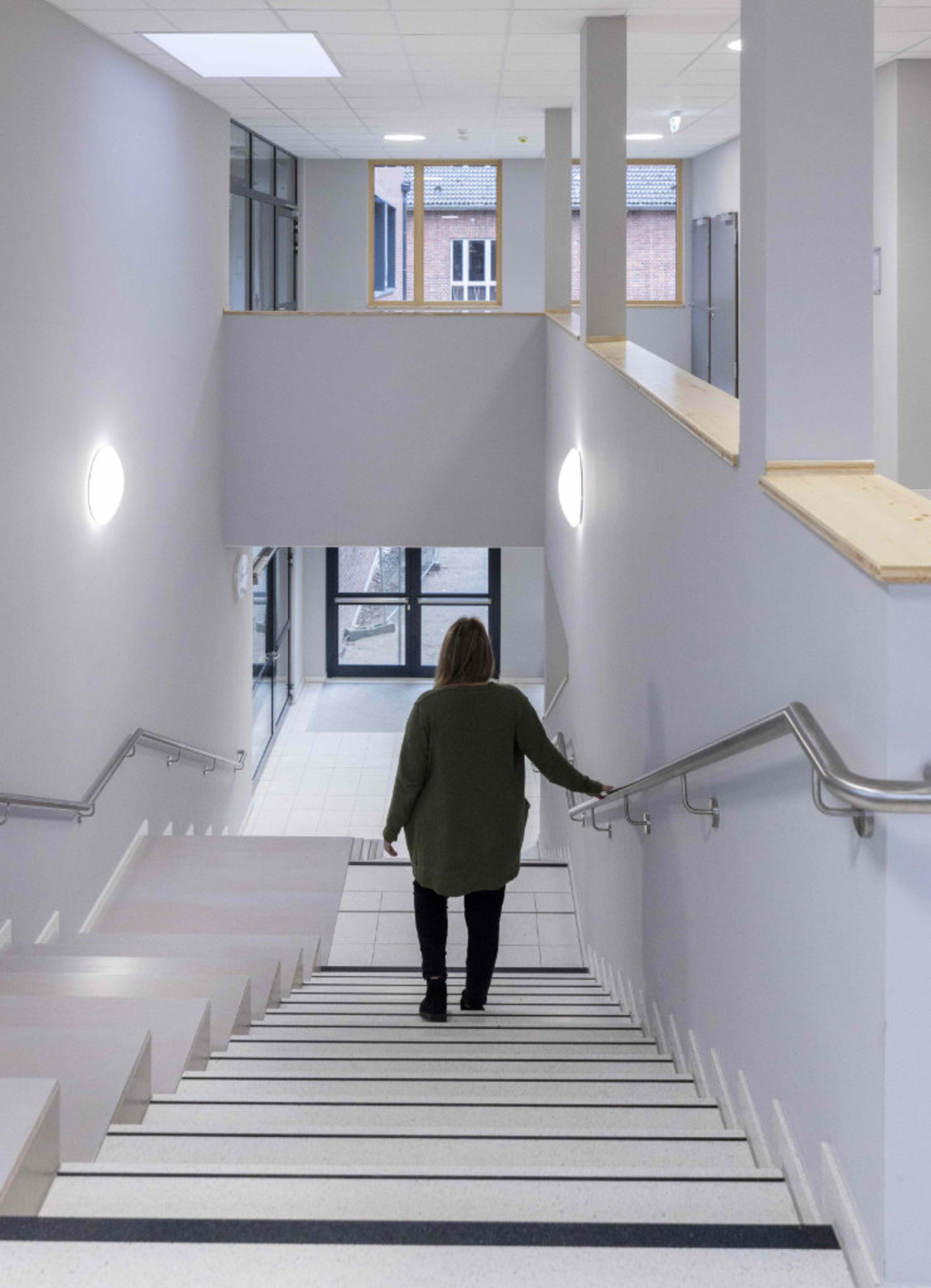 Eine Frau geht im Modulbau durch den hellen Flur vom Obergeschoss in das Erdgeschoss. Durch die Fenster mit Holzrahmen ist der Altbau zu sehen. Der Nebau hat wie er eine Klinkerfassade erhalten.