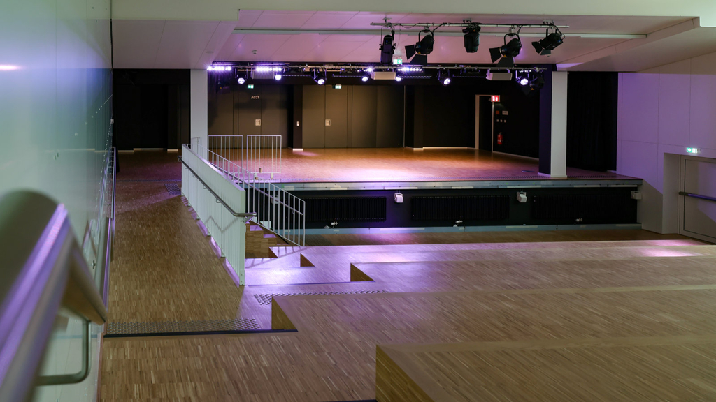 Blick in die noch unbestuhlte Aula mit Parkett wie in Terrassen hinunter bis zur Bühne, die von Scheinwerfern an der Decke angestrahlt wird.