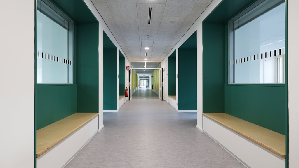 Ein langer, heller Flur, der immer wieder von Scheiben mit Blick in die Klassenzimmern unterbrochen wird. Vor den Scheiben befinden sich Sitznischen, die Wände dazu sind in einem dunklen Grün gestrichen.