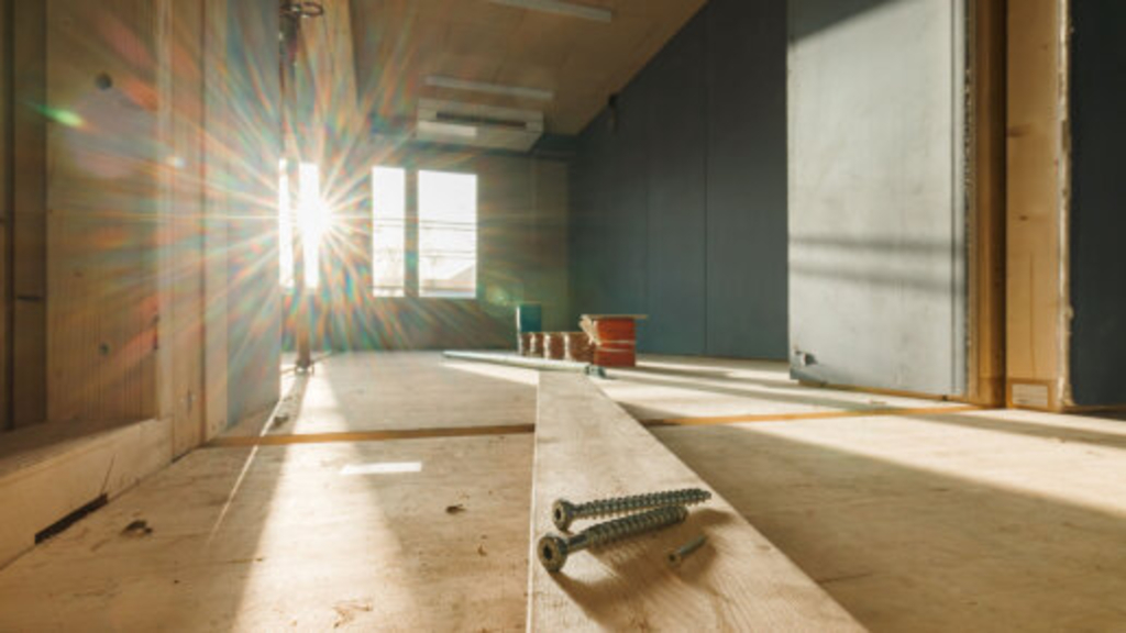 Drei Schrauben auf einer Holzdiele liegend, die in einen künftigen Klassenraum reicht. Durch das Fenster dahinter ist strahl die Sonne herein., Link auf Holzbauten – von Natur aus gut - Stadt Köln