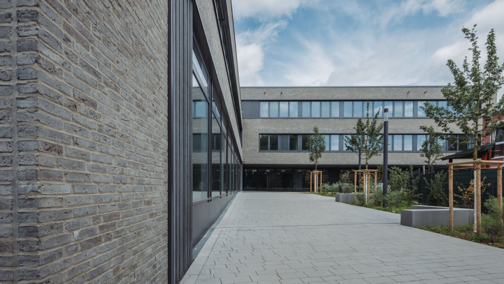 Eine grau verklinkerte Fassade, eine Fensteranlage dahinter und daneben ein gepflasterter Weg mit neu gepflanzten Bäumen, der zum Haupteingang führt.
