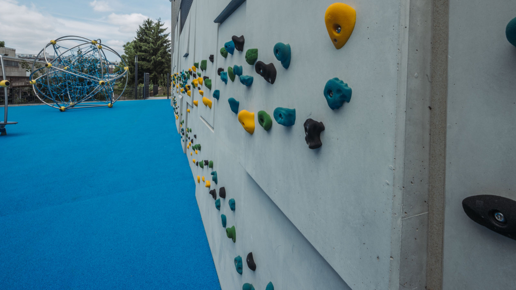 Ein blauer Bodenbelag mit Klettergerüst aus Tauen an der hinteren Rückseite des unteren Schulhof. An der Fassade ebenfalls eine Kletteranlage mit kleinen, bunten Elementen, die an der Wand befestigt als Auflagefläche für die Füße dienen.
