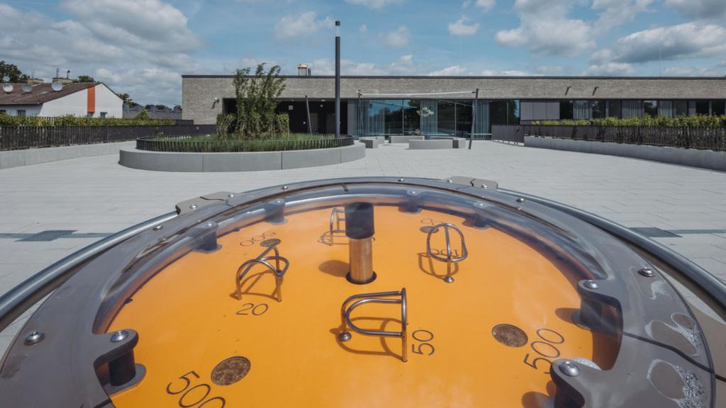 Auf dem Schul-Dachgarten ist im Vordergrund eine sonnengelbes, rundes Geschicklichkeitsgerät zu sehen, auf dem Kugeln in Mulden mit Punktzahlen zu manövrieren sind. Dahinter grüne Heckenpflanzen und junge Bäume und Sträucher mit Sitzgelegenheiten und Sonnensegel. 