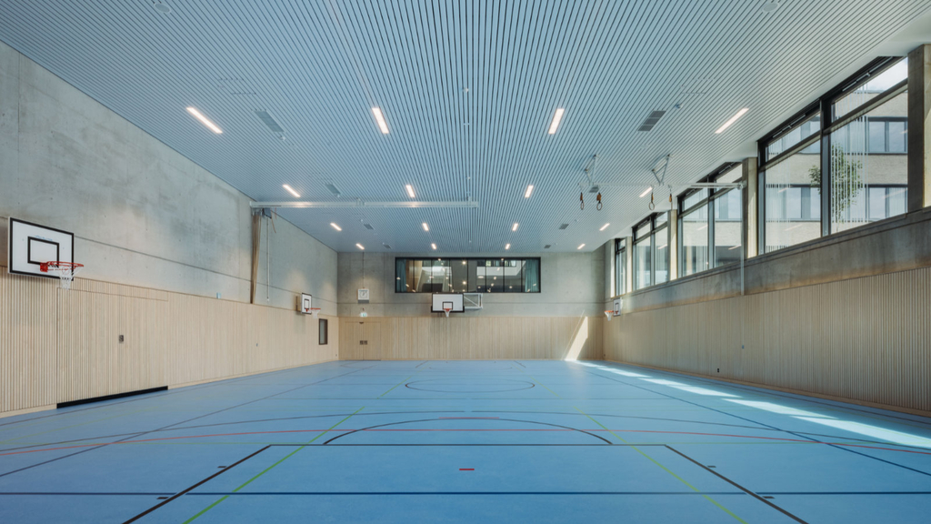 Die Sporthalle mit blauem Bodenbelag und Spielfeldmarkieren, Basketballkörben in verschiedenen Höhen sowie Fenstern mit innenliegendem Sonnenschutz.