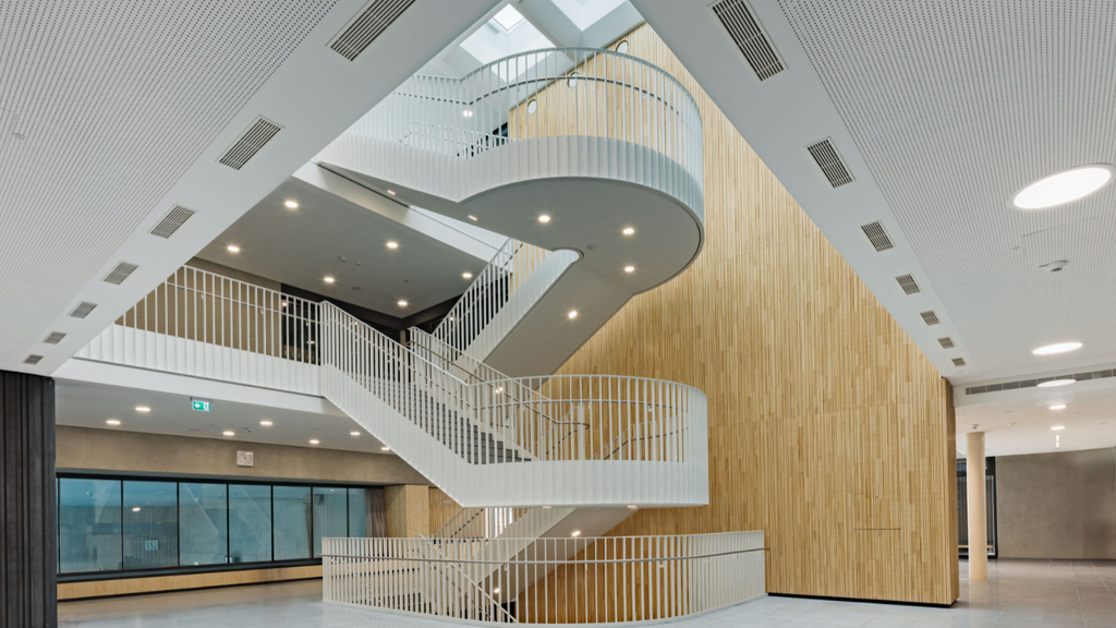 Durch ein Atrium fällt sehr viel Tageslicht in ein Foyer mit offenem Treppenhaus, das an den Wänden mit hellem Holz verkleidet wurde. Dahinter eine Sitznische mit Fensterreihe, die Blicke in die darunter liegende Sporthalle ermöglicht.