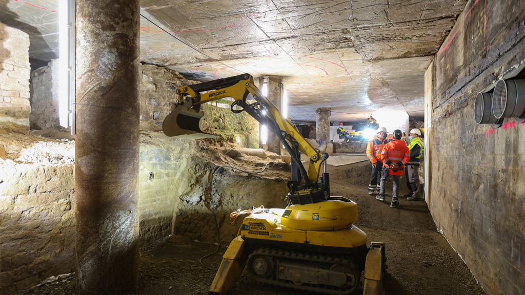 Ein Minibagger unter Tage in der Archäologischen Zone. Im Hintergrund stehen drei Arbeiter und besprechen sich.