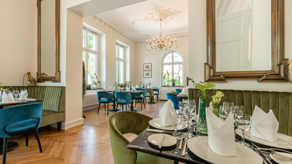 Innenansicht der Villa Bodinus. Zu sehen sind festlich eingedeckte Tische an grünen und blauen Stühlen. Im hinteren Bereich hängt ein Kronleuchter unter der Decke und an den Wänden sind Spiegel aufgehängt.