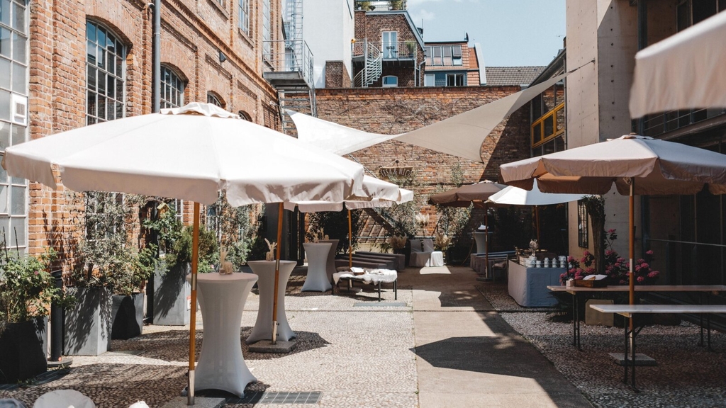 Außenbereich der KunstSalon Location. Zu sehen sind aufgestellte Sonnenschirme und Stehtische mit Hussen.