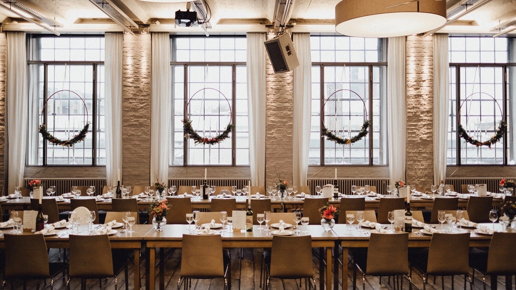 Festlich gedeckter langer Holztisch, mit davorstehenden Holzstühlen. Im Hintergrund sind vier große Fenster  zu sehen, die mit geschmückten Metallringen dekoriert sind. 