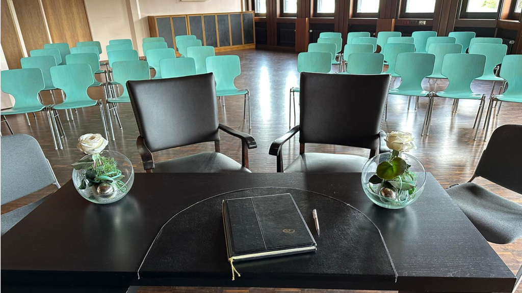 Innenansicht des Trauzimmers im Bürgeramt Porz. Im Vordergrund steht ein Trautisch in dunklem Holz, im Hintergrund stehen türkisfarbene Stühle für die Gäste.