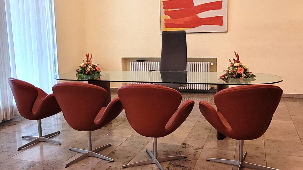 Trauzimmer im Rathaus - Spanischer Bau: abgebildet ist ein ovaler, mit Blumen geschmückter gläserner Trautisch mit Stühlen vor einem bunten Gemälde.