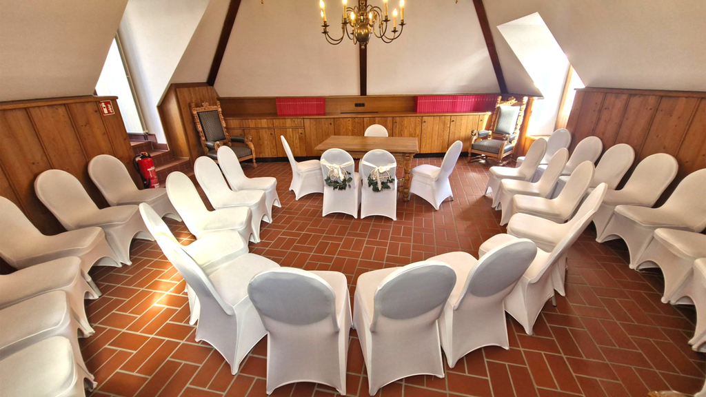 Trauzimmer Turm: In der hinteren Mitte des Raums befindet sich unter einem Kronleuchter ein Trautisch mit vier Stühlen für das Hochzeitspaar und Trauzeugen. Im Halbkreis hinter dem Hochzeitspaar sind zwei Reihen von Stühlen für Gäste platziert. Alle Stühle sind mit weißen Hussen geschmückt.