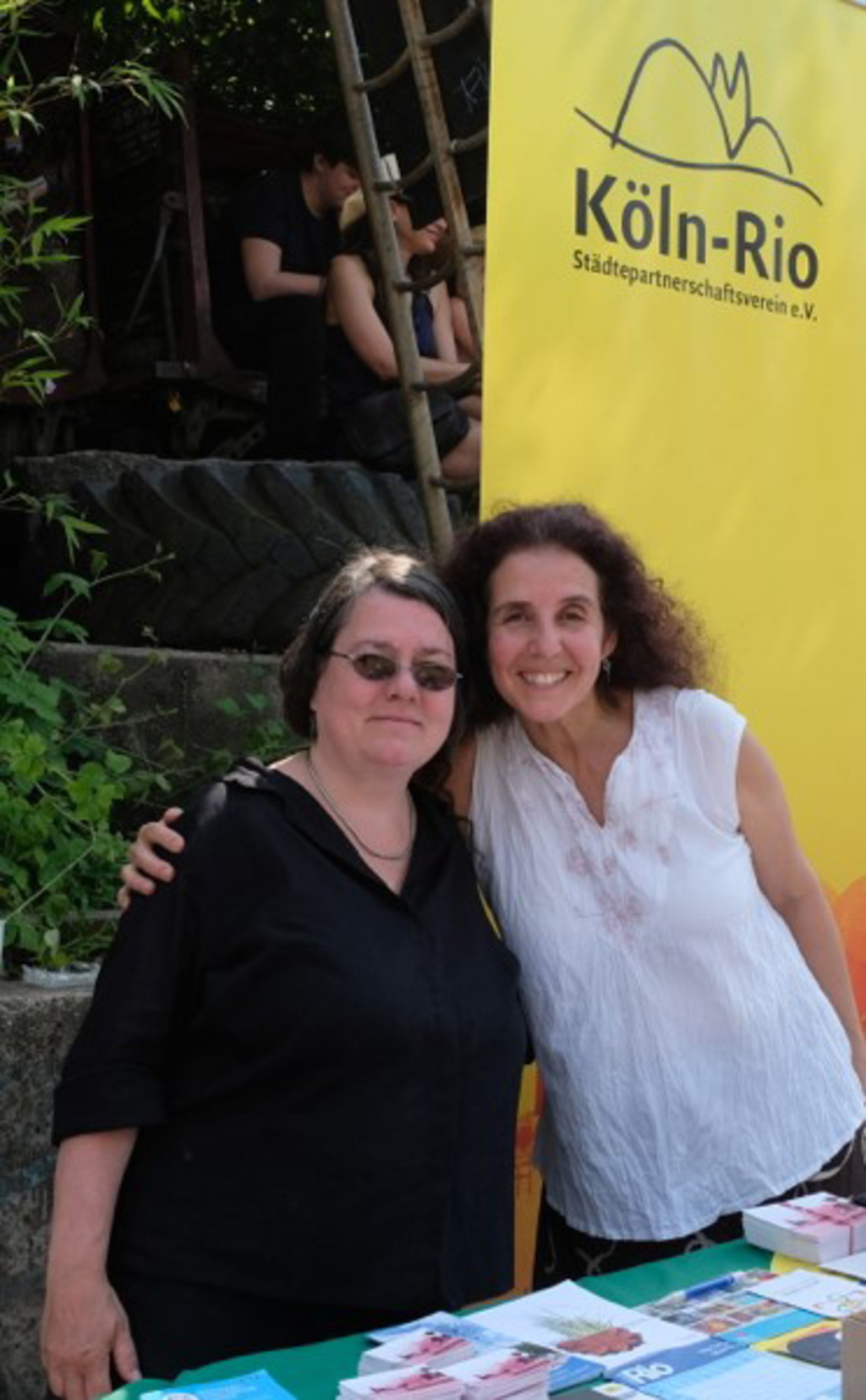 Zwei Frauen stehen draußen vor einem Köln-Rio-Banner und lächeln in die Kamera