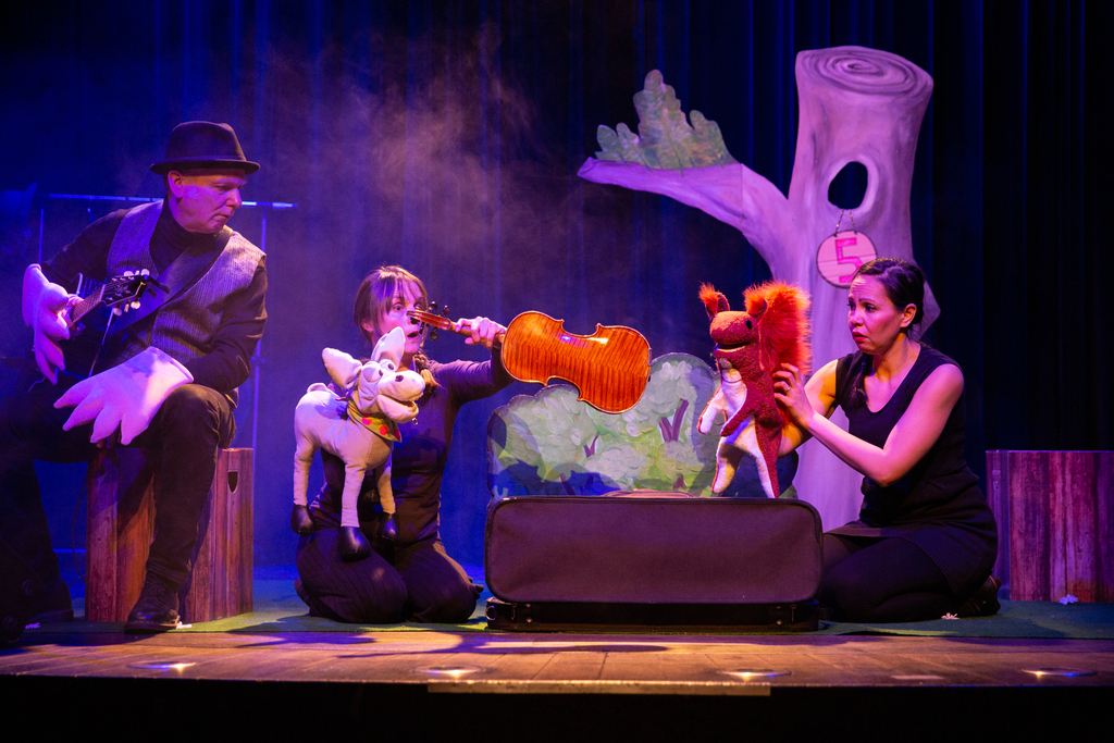 Vor einem Baum-Bühnenbild sitzen 3 Schauspieler*innen mit ihren Handpuppen und spielen eine Szene