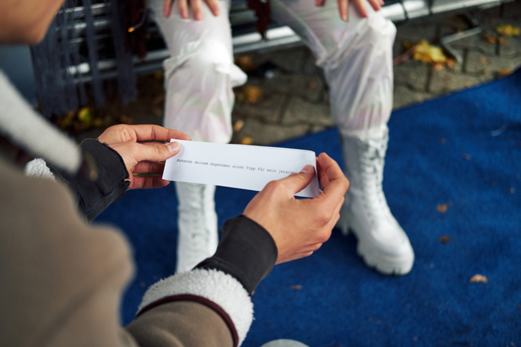 Hände halten einen beschrifteten Zettel, im Hintergrund sind Beine zu sehen