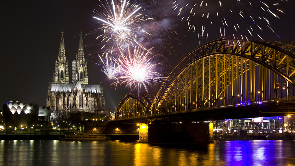 Silvesterfeuerwerk in Köln. Zu sehen sind mehrere bunte Feuerwerkraketen. Beleuchtet ist der Kölner Dom und die Hohenzollernbrücke.