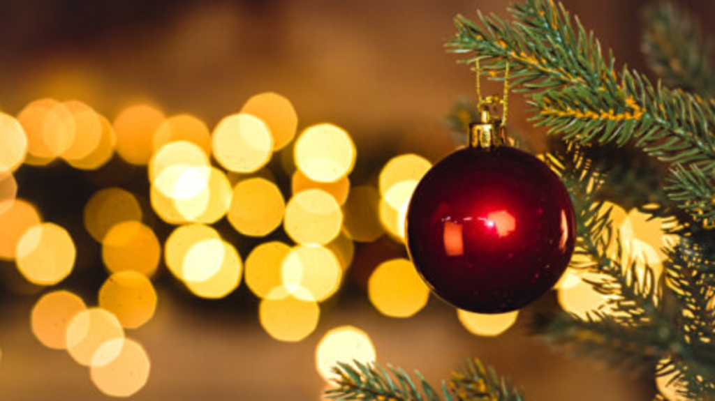 Eine rote Weihnachtskugel hängt an einem Tannenzweig. Im Hintergrund schimmern Lichter., Link auf Geschenktipps - Stadt Köln