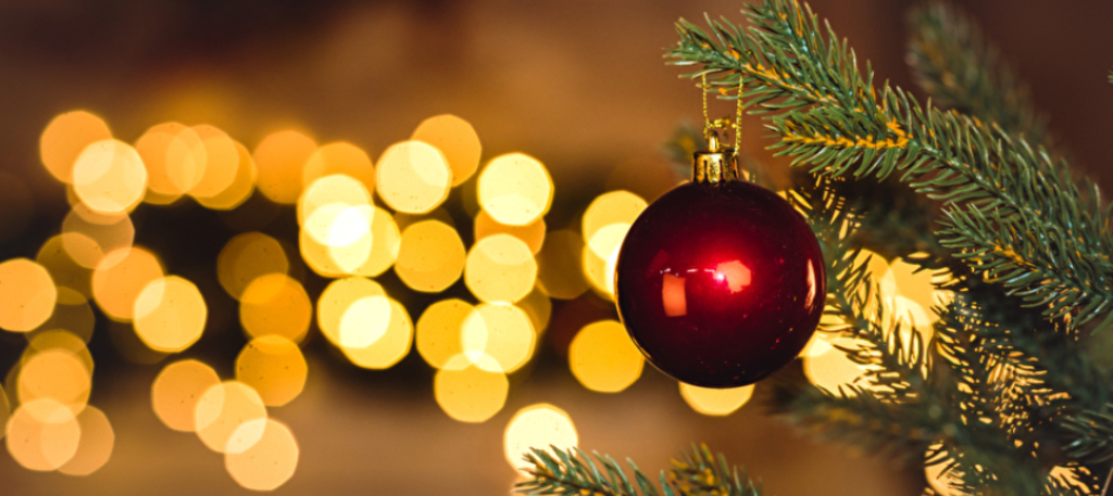 Eine rote Weihnachtskugel hängt an einem Tannenzweig. Im Hintergrund schimmern Lichter.