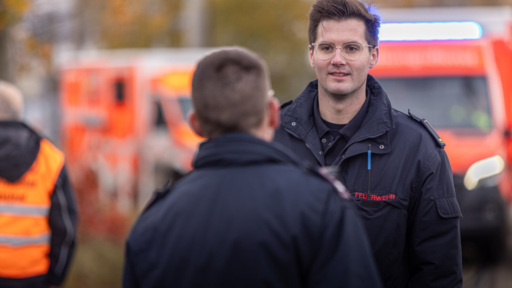 Zwei Feuerwehrkräfte unterhalten sich. Im Hintergrund sind Einsatzfahrzeuge zu sehen., Link auf Ausbildung als Brandreferendar*in