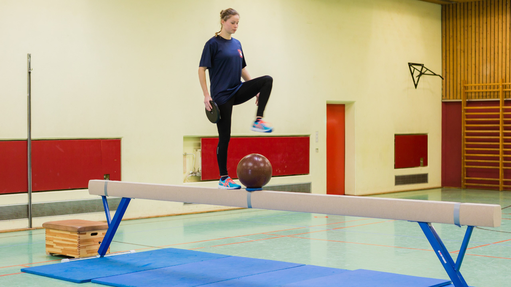 Frau balanciert mit Gewicht in der Hand über einen Schwebebalken, in dessen Mitte ein Medizinball liegt., Link auf Eignungstest für Brandmeister*innen und Stufenausbildung Brandmeister*innen