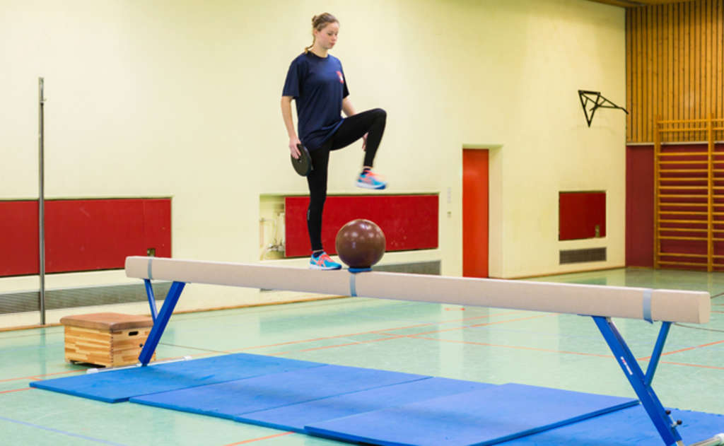 Frau balanciert mit Gewicht in der Hand über einen Schwebebalken, in dessen Mitte ein Medizinball liegt.