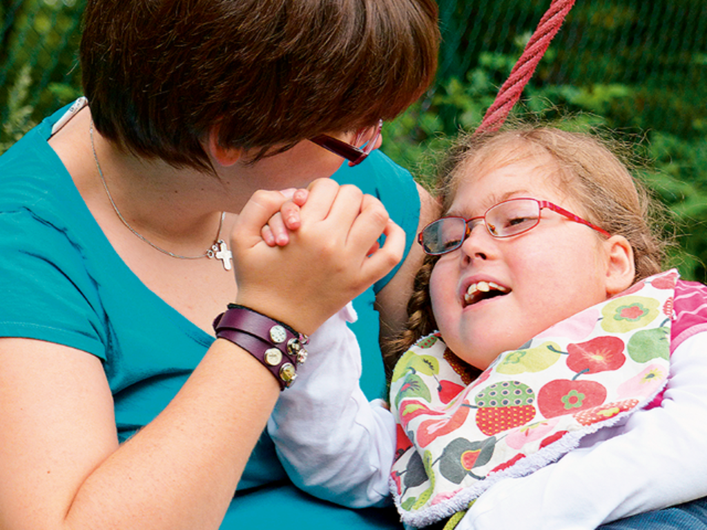 Frau wiegt ein pflegebedürftiges junges Mädchen im Arm und hält dessen Hand