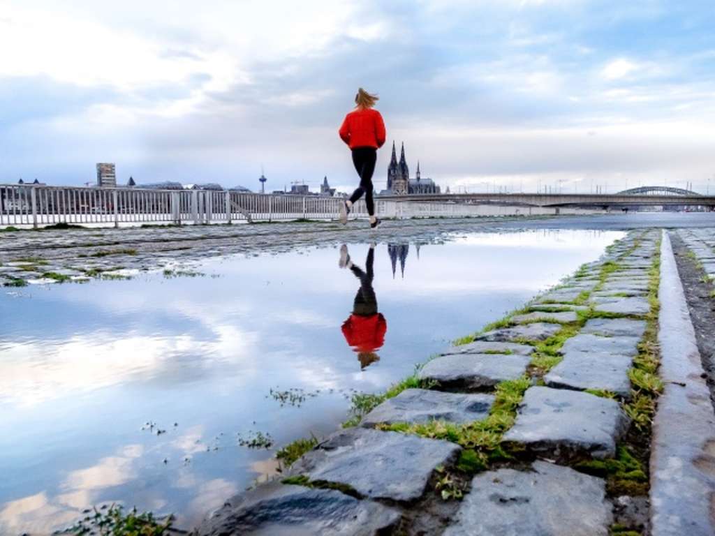 Frau joggt am Rheinufer entlang, die Kölner Skyline im Hindergrund