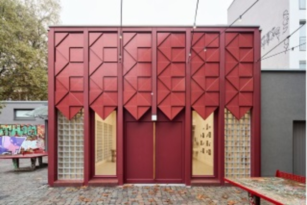 Das Foto zeigt den Eingang in die Holzwerkstatt der Alten Feuerwache Köln. Der untere Teil des Eingangs besteht ein aus einer geschlossenen Doppeltür, eingerahmt von Glastüren. Der obere Teil des Eingang ist mit einem Rautenmuster in bordeauxrot verkleidet.