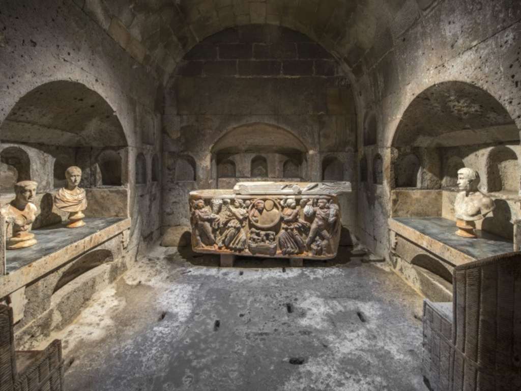 Ein Blick in die Römergrabkammer Köln Weiden, mittig ein reichlich verzierter Sarg aus der Antike, an den seitlichen Auswölbungen des Raumes antike Büsten.