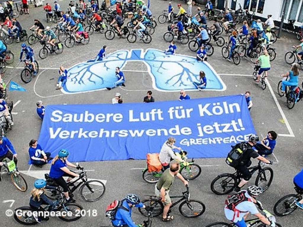 Fahrrad-Demo: Unzählige Fahrradfahrende umkreisen ein riesiges Plakat "Saubere Luft für Köln, Verkehrswende jetzt! Greenpeace" und eine aufgemalte blaue Lunge.
