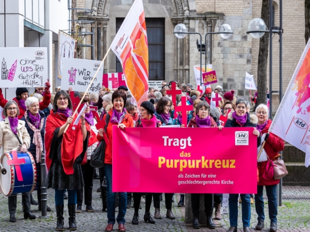 Frauengruppe mit Fahnen vor dem Kölner Dom, sie halten ein Banner mit der Aufschrift "Tragt das Purpurkreuz"