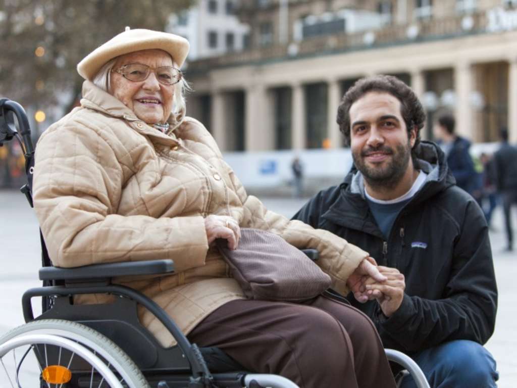 Eine Rentnerin im Rollstuhl, neben Ihr hockt ein Ehrenamtlicher und hält Ihre Hand. Beide schauen lächend in die Kamera.