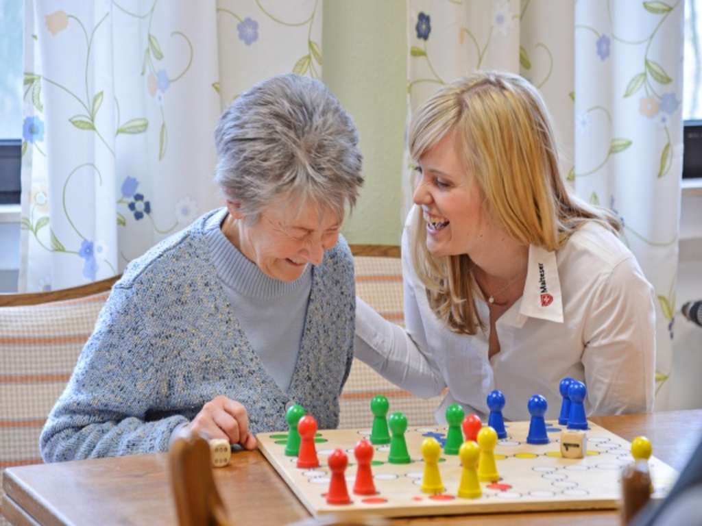 Fröhlicher Umgang zwischen junger Frau der Malteser und Seniorin, die zusammen Mensch ärgere dich nicht spielen
