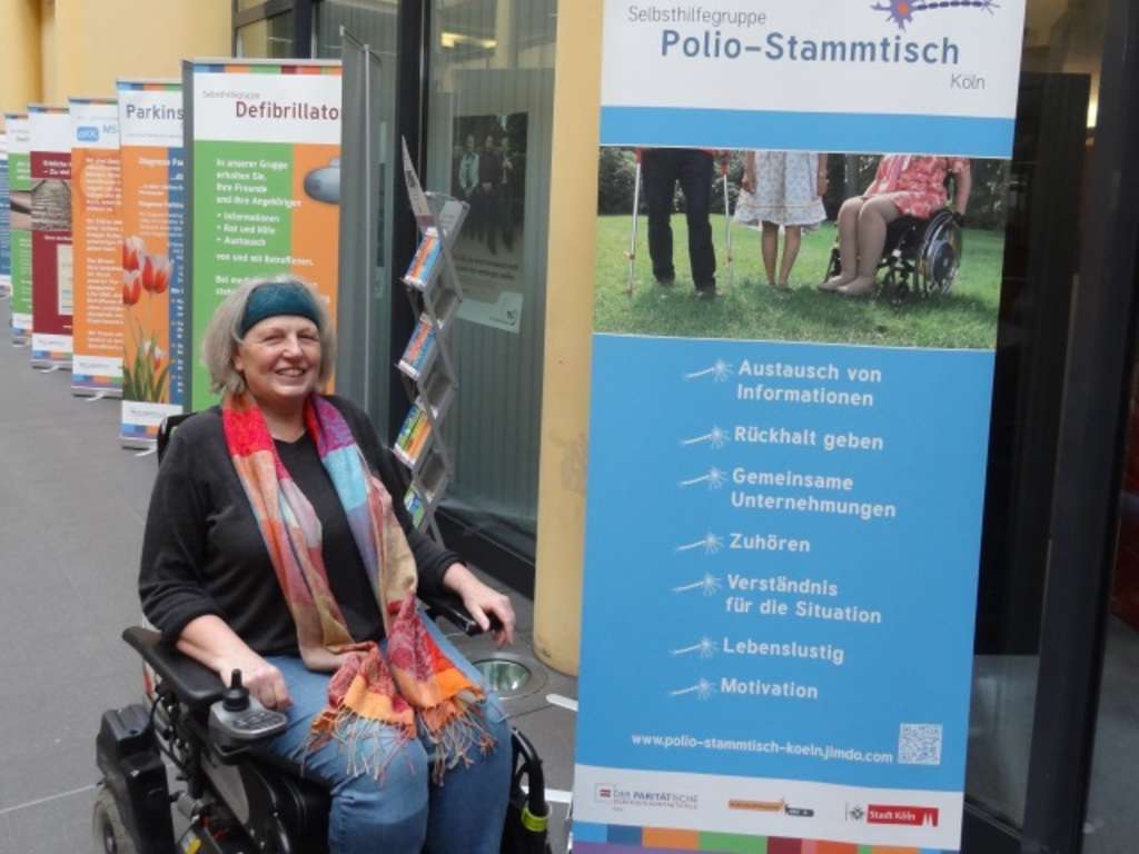 Frau im Rollstuhl neben einem Roll up der Selbsthilfegruppe Polio-Stammtisch Köln