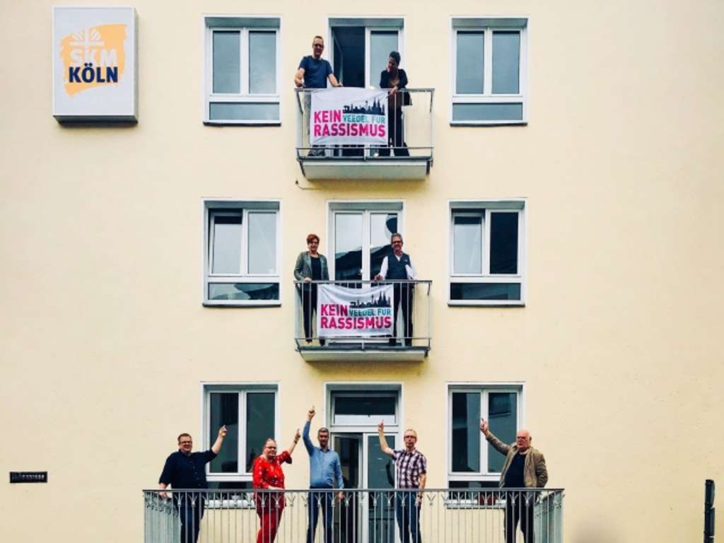 Mehrstöckiges Wohnhaus, auf den Balkonen hängen Banner "Kein Veedel für Rassismus"