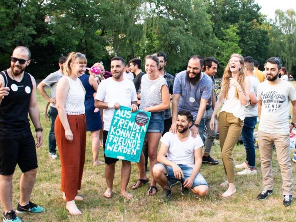 Junge, interkulturelle Menschen stehen lachend zusammen, wobei einer ein Plakat in der Hand hält mit der Aufschrift "Aus Fremden können Freunde werden"