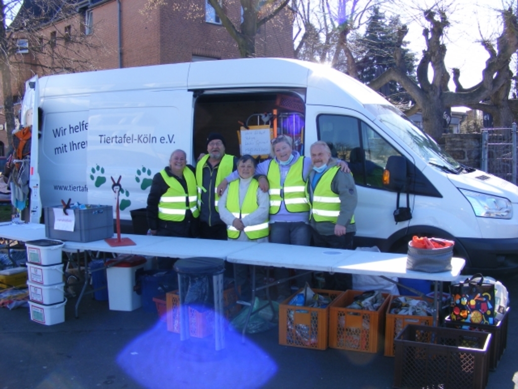 Fünf Ehrenamtliche bei der Ausgabe von Tierbedarf. Im Vordergrund mehrere gefüllte Kisten, im Hintergrund der weiße Transportwagen mit der Aufschrift Tiertafel-Köln e. V. 