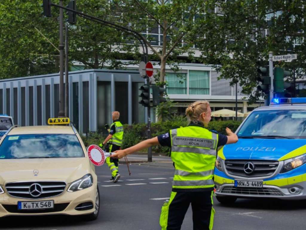 eine Verkehrskadettin, zu erkennen an ihrer Signalkleidung und einer sogenannten Verkehrskelle in der Hand, steht auf der Kreuzung und regelt den Verkehr. Im Hintergund ist ein Polizeibus zu sehen.