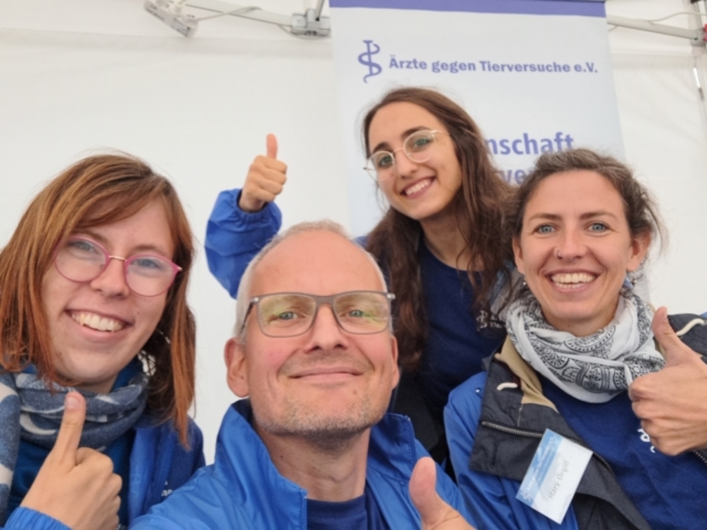 Vier Ehrenamtliche der Arbeitsgruppe Köln von Ärzte gegen Tierversuche e. V. stehen vor einem Infostand. Alle lächeln und zeigen den Daumen hoch.