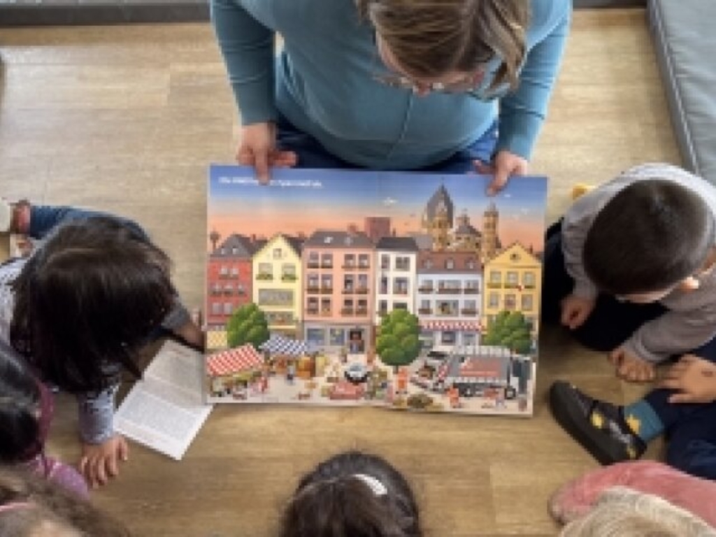 Sechs Kindern im Grundschulalter wird aus einem Bilderbuch vorgelesen, das eine erwachsene Frau mit Blickrichtung zu den Kindern vor sich hält. Das Bilderbuch klärt auf zum Thema „Müllvermeidung“.