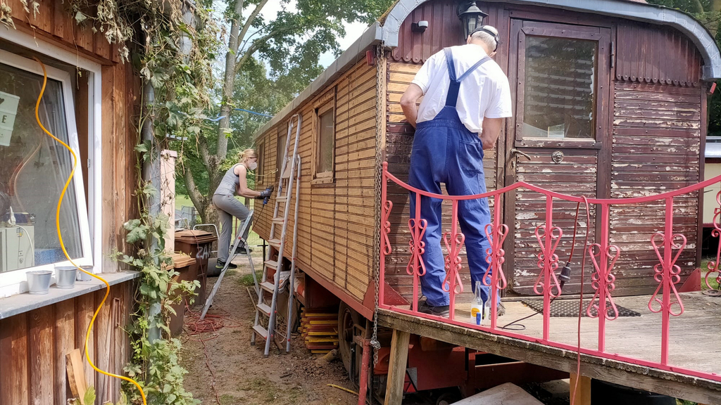 Im Vordergrund ein Mann, den Rücken zugekehrt, bei der Arbeit am historischen Zirkuswagen. Im hinteren Bereich des Bildes steht eine Frau mit Mundschutz auf einer Leiter, in der Hand ein Schleifgerät, mit dem sie gerade die Holzfassade des Zirkuswagens schleift.