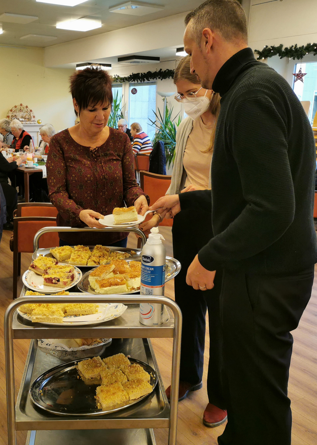 : Zahlreiche Streusel-Kuchenstücken auf einem Servierwagen, daneben drei Personen bei der Verteilung auf Teller. Im Hintergrund sitzen Senior*innen an einem gedeckten Tisch.